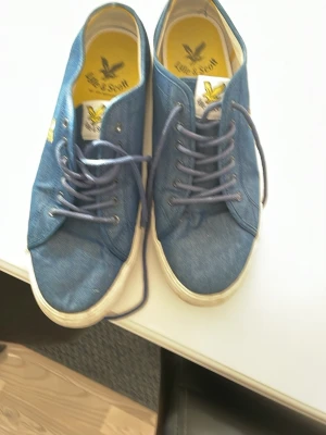 Blåa seglarskor från Lyle & Scott - Säljer ett par blåa seglarskor från Lyle & Scott med vita sulor och klassisk snörning. Skorna har en gul logga på innersulan och bruna yttersulor med hexagonmönster. Materialet är tyg och de har en avslappnad, stilren look.