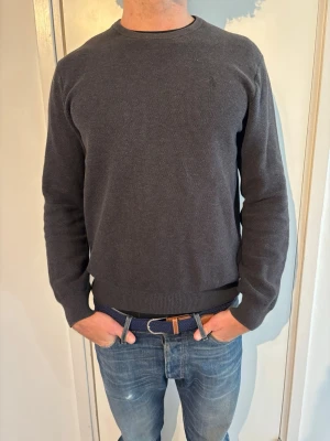 Polo Ralph Lauren tröja   - Klassisk mörkgrå tröja från Polo Ralph Lauren med rund halsringning och ribbade muddar vid ärmslut och nederkant. Tröjan är enkel och stilren, perfekt till jeans eller chinos. Materialet är mjukt och bekvämt, vilket gör den skön att bära hela dagen.