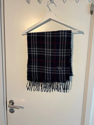 Burberry halsduk - Snygg halsduk från Burberry i klassiskt rutigt mönster med svart, vitt, grått och röda detaljer. Tillverkad i 100% lammull och har fransar i kanterna. Perfekt accessoar för att lyfta din outfit med en tidlös brittisk touch.