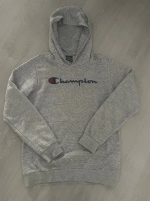 Grå Champion hoodie med logga - Säljer en klassisk grå hoodie från Champion med stor broderad logga i marinblått och rött över bröstet. Hoodien har huva, magficka och ribbade muddar vid ärmar och nederkant. Tillverkad i mjuk bomullsblandning. (13/14 år)