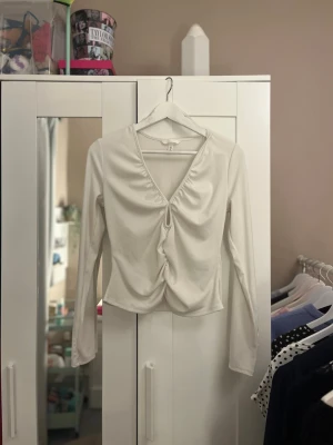Vit omlottopp med rynk framtill - Trendig vit topp med v-ringning och snygg rynkdetalj framtill. Modellen är croppad och har långa ärmar. Perfekt för dig som gillar stilrena plagg med en twist. Tillverkad i mjukt material som känns skönt mot huden.