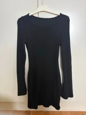 Svart ribbad långärmad klänning - En svart, ribbad kortklänning med lång ärm och rund halsringning. Klänningen har en kroppsnära passform och är tillverkad i ett stretchigt material som ger en bekväm känsla. Perfekt för dig som gillar en enkel men stilren look.