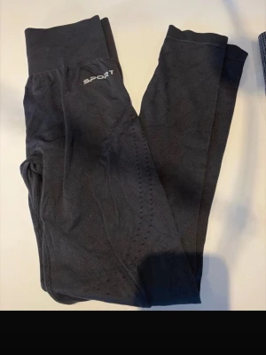 Svarta träningstights med hög midja - Säljer ett par svarta träningstights från Sport med hög midja och diskret logga på höften. Tightsen är stretchiga och har ventilerande detaljer längs benen, perfekta för gymmet eller löprundan.