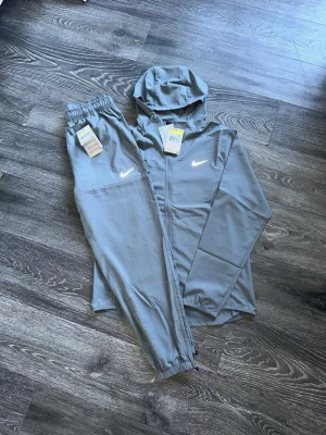 Grått Nike träningsset med huva-  M/S junior  - Snyggt grått träningsset från Nike med matchande byxor och zip-hoodie. Setet har vita Nike-loggor på både jackan och byxan. Jackan har huva och dragkedja, byxorna har resår i midjan och vid benslut. Perfekt för gymmet eller löprundan.