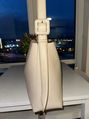 Beige axelväska med justerbart band - Stilren beige axelväska i slätt konstläder med fyrkantig form och bred axelrem med stor spänndetalj. Väskan har minimalistisk design och rymligt huvudfack. Har lite fläckar som går att ta bort samt slitning se bilder. Detta ser man inte om man ej kollar super noga.