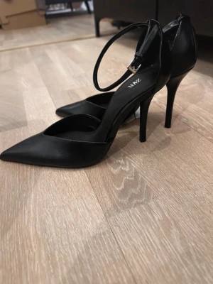 Svarta klackar  Zara - Säljer ett par svarta pumps från Zara med spetsig tå och smal klack. Skorna har en elegant ankelrem med silverfärgat spänne och är tillverkade i slätt konstläder. Perfekta för att lyfta vilken outfit som helst.