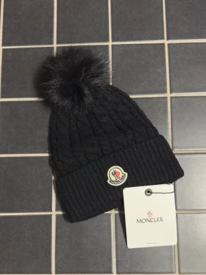 Svart Moncler mössa med pälsboll - Snygg svart stickad Moncler mössa med stor pälsboll på toppen och Moncler-logga framtill. Mössan har bred uppvikt kant och kabelstickat mönster som ger en exklusiv känsla. Perfekt accessoar för kalla dagar.