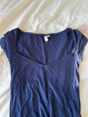 Mörkblå topp med spetskant från Nelly - Snygg mörkblå topp från Nelly med v-ringning och dekorativ spetskant runt halsen. Tillverkad i mjuk bomull och har korta ärmar. Perfekt att styla med jeans eller kjol för en trendig look. Aldrig använd !! 