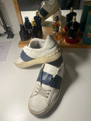 Valentino vita sneakers med blå detalj - Snygga vita sneakers från Valentino med bred blå remsa över mitten och coola nitar längs sulan. Skorna har en klassisk låg profil och är tillverkade i läder. Perfekta för dig som vill ha en stilren men ändå unik look.