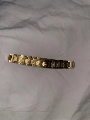 Lacoste armband  - Snyggt guldigt armband med breda länkar i metall. Armbandet har en rektangulär form på länkarna och ett diskret mönster på varje länk som ger en cool och unik look. Perfekt accessoar för att lyfta din outfit.