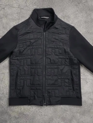 Svart quiltad jacka från J.Lindeberg - Säljer en svart jacka med quiltat mönster på framsidan och axlarna. Jackan har ståkrage, dragkedja framtill och två sidofickor med dragkedja. Materialet är en mix av slät textil och vadderad panel. Perfekt för dig som gillar stilren och modern look.