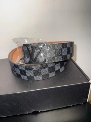 LV Bälte - Snyggt LV-bälte med klassiskt rutigt mönster i svart och grått. Bältet har en stor LV-logga i metall som spänne och insidan är i ljust skinn. Perfekt accessoar för att lyfta din outfit.110CM verktyg för att justera storlek följer med 
