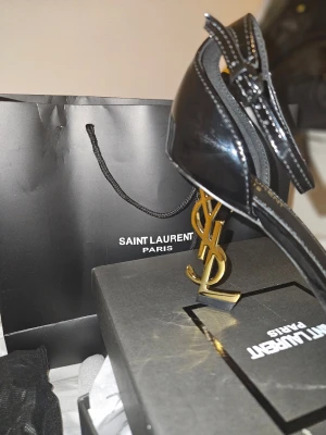 Svarta Saint Laurent sandaler - Säljer ett par svarta sandaler från Saint Laurent i lackat läder med öppen tå och fyrkantig framdel. Klackarna har den ikoniska YSL-loggan i guld som ger en lyxig touch. Justerbart ankelband med spänne. Perfekta för dig som vill sticka ut.