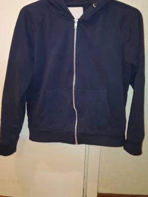 Svart hoodie med dragkedja - En enkel svart hoodie med dragkedja framtill och huva. Modellen har långa ärmar, breda muddar och en klassisk passform. Perfekt för en avslappnad stil och funkar till det mesta. Materialet känns mjukt och bekvämt, troligen bomullsblandning.