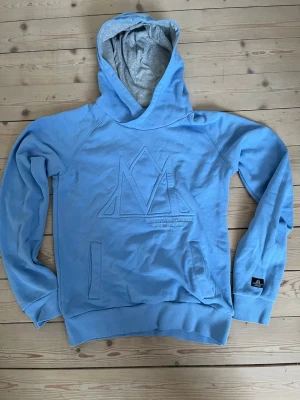 Blå hoodie från Mountain Horse - Ljusblå hoodie från Mountain Horse med stor broderad logga på bröstet och känguruficka framtill. Huvan är fodrad i grått och muddar finns vid ärmslut och nederkant. Perfekt för en chill och sportig stil.