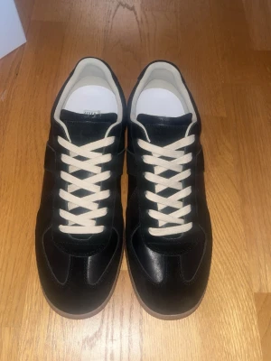 Svarta maison margielas - Snygga svarta sneakers med vita snören och beige sula. Skorna har en klassisk låg siluett och är tillverkade i en mix av läder och mocka. Insidan är ljus och ger en fräsch kontrast till det svarta yttermaterialet. Perfekta för en clean och stilren look. Box ingår med annat. Skriv om frågor storlek 43