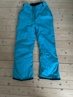 Turkosa täckbyxor från Everest - Säljer ett par turkosa täckbyxor från Everest med raka ben och dragkedjor vid fickorna. Byxorna har breda hällor för bälte och är perfekta för kalla dagar i backen. Materialet är slitstarkt och vattenavvisande, vilket gör dem grymma för vinterns alla äventyr.