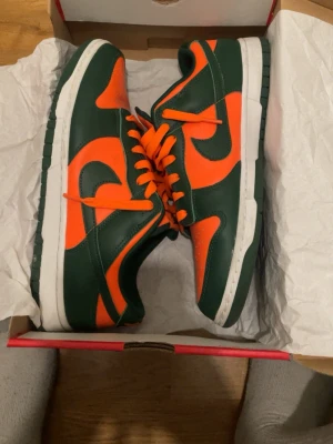 Nike Dunk Low grön/orange sneakers - Nike Dunk Low sneakers i grön och orange färg med klassisk vit sula. Skorna har en låg siluett, orange swoosh och matchande orange skosnören. Tillverkade i läder med en robust yttersula för bra grepp. Perfekt för dig som vill sticka ut med färg på fötterna.