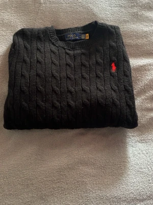 Svart kabelstickad tröja Polo Ralph Lauren - Kabelstickad svart tröja från Polo Ralph Lauren med rund halsringning och röd broderad logga på bröstet. Tröjan har ribbade muddar vid ärmslut och nederkant och är tillverkad i mjukt material. Perfekt för en stilren och klassisk look.