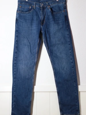 Levi's blå raka jeans - Klassiska blå jeans från Levi's med rak passform och fem fickor. Jeansen har en djupblå tvätt, gylf med dragkedja och knapp samt detaljerade sömmar. Tillverkade i slitstarkt denim med den ikoniska läderpatchen bak i midjan.