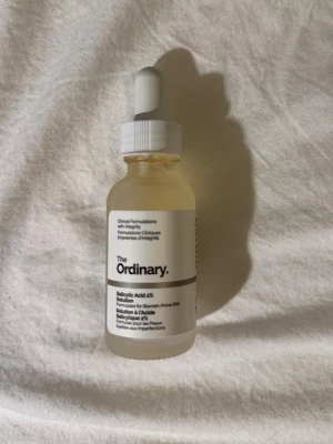 The Ordinary Salicylic Acid 2% Solution - Salicylsyra 2% lösning från The Ordinary i en genomskinlig glasflaska med pipett. Perfekt för dig som vill behandla orenheter och finnar. Flaskan har vit etikett och vitt lock. Enkel att använda i din hudvårdsrutin.