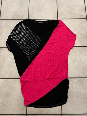 Svart och rosa topp med nitar storlek S/M - Cool topp från K-Klass i svart och starkt rosa med diagonal färgdelning. Ena axeln är dekorerad med silvriga nitar som ger extra edge. Toppen har v-ringning och är kortärmad med loose passform, perfekt för en trendig look.