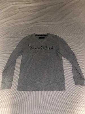 Grå sweatshirt från Bondelid - Snygg grå sweatshirt från Bondelid med rund halsringning och svart broderad logga på bröstet. Tröjan har långa ärmar och ribbade muddar vid ärmslut och nederkant. Perfekt för en avslappnad och stilren look. Tröjan har dock två små hål vid varsin armbåge.