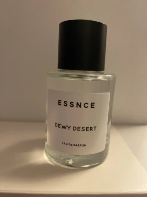 ESSNCE Dewy Desert Eau de Parfum - Minimalistisk parfymflaska från ESSNCE med doften Dewy Desert. Flaskan är cylindrisk i genomskinligt glas med en svart, rund kork och vit etikett med svart text. Modern och stilren design som passar perfekt på parfymhyllan. 4 sprut tagna 