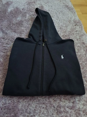 Svart hoodie från Polo Ralph Lauren - Klassisk svart hoodie från Polo Ralph Lauren med dragkedja framtill och vit broderad logga på bröstet. Hoodien har justerbar huva med snören och är tillverkad i mjukt bomullsmaterial. Perfekt för en chill och stilren look. Lite noppig och urtvättad, därav det fina priset! Öppen för förhandling! Skriv vid funderingar!