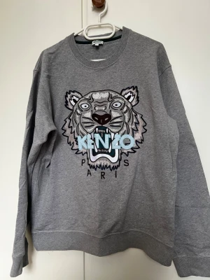 Grå Kenzo sweatshirt  - Säljer en grå sweatshirt från Kenzo med det ikoniska tigerhuvudet broderat framtill och texten 'KENZO PARIS' i blått och svart. Tröjan har rund halsringning, ribbade muddar och är tillverkad i mjukt bomullsmaterial.