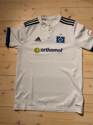 Vit HSV matchtröja från Adidas - Säljer en vit matchtröja från Hamburger SV med blå och svarta detaljer. Tröjan har klubbmärke, Adidas-logga och Orthomol som sponsortryck på bröstet. Korta ärmar med blå kant och svart axelrand. Tillverkad i funktionsmaterial, perfekt för fotbollsträning.