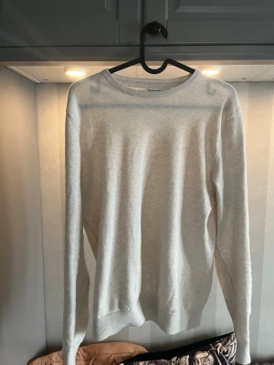 Ljusgrå basic tröja rund hals - En stilren ljusgrå tröja med rund halsringning och långa ärmar. Tröjan har en enkel, klassisk design med ribbade muddar vid ärmslut och nederkant. Perfekt att matcha med jeans eller kjol för en clean look.