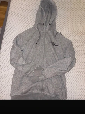 Grå zip hoodie från Peak Performance - Snygg grå hoodie från Peak Performance med dragkedja framtill och broderad logga på bröstet. Tröjan har huva med dragsko och ribbade muddar vid ärmslut och nederkant. Perfekt för en avslappnad och sportig stil. Tyvärr är det ett litet hål som syns på bilden