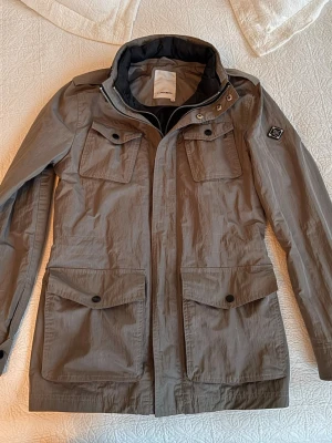 Grå Jacka från J.Lindeberg - Snygg grå field jacket från J.Lindeberg med fyra stora fickor framtill och hög krage. Jackan har en diskret logga på ärmen och stängs med dragkedja och knappar. Perfekt för kyliga dagar och har en stilren, modern look.