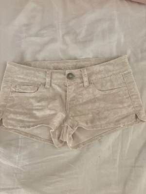 Beige mönstrade shorts från american eagle - Säljer ett par ljusbeige shorts från american eagle med lite mönster. Modellen är lågmidjad med klassiska fickor fram och bak, bälteshällor och knappstängning. Perfekta för varma sommardagar och har en snygg, tight passform.