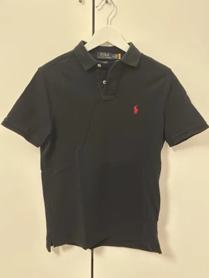Svart pikétröja från Polo Ralph Lauren - Klassisk svart pikétröja från Polo Ralph Lauren med korta ärmar och krage. Tröjan har två knappar vid halsen och den ikoniska röda logotypen broderad på bröstet. Tillverkad i mjuk bomull för en bekväm känsla.