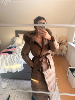 Lång brun dunjacka med pälskrage - Snygg lång brun dunjacka med bälte i midjan och stor huva med fluffig fuskpälskant. Jackan har en figurnära passform och knäpps med knappar framtill. Perfekt för kalla vinterdagar och ger en trendig siluett.