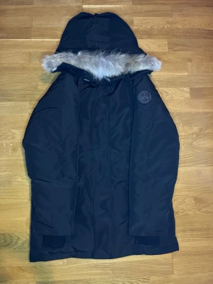 Svart Canada Goose parkas med päls - Säljer en svart parkas från Canada Goose med huva och snygg pälsdetalj. Jackan har ett broderat märke på ärmen och dragkedja framtill. Perfekt för kalla vinterdagar och har en klassisk, clean look. Medföljer originalpåse från Canada Goose.