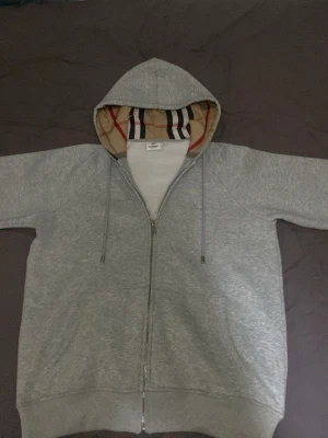 Grå zip-hoodie från Burberry - Snygg grå hoodie med dragkedja från Burberry. Hoodien har klassiskt rutigt foder i huvan, dragsnören och två fickor framtill. Perfekt för en chill look.