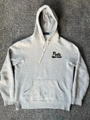 Grå hoodie från Polo Ralph Lauren - Snygg ljusgrå hoodie från Polo Ralph Lauren med svart broderad Polo-logga och texten 'Est. 1967' på bröstet. Klassisk känguruficka framtill och justerbar dragsko i huvan. Tillverkad i mjuk bomullsblandning, perfekt för chill dagar.