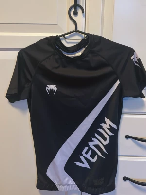 Svart rashguard från Venum - Säljer en svart kortärmad rashguard från Venum med vit logga och grafiskt tryck på framsidan. Tröjan har en tight passform och är tillverkad i ett stretchigt funktionsmaterial som passar perfekt för kampsport eller intensiv träning.