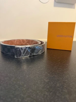 L V Damier bälte grå - Snyggt L.V bälte i klassiskt Damier-mönster i grå toner. Bältet har ett stort L.V-spänne i silverfärg och kommer med originalförpackning och kvitto. Perfekt accessoar för att lyfta din outfit.