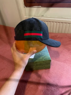 Svart Gucci keps med röd/grön rand - Snygg svart keps från Gucci med klassiskt GG-mönster över hela kepsen och en ikonisk grön och röd rand på sidan. Kepsen har böjd skärm och justerbar passform. Materialet ser ut att vara bomullscanvas med exklusiv känsla. Perfekt för dig som vill ha en stilren och lyxig accessoar.