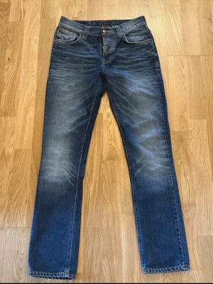 Blå jeans från Nudie Jeans - Snygga blå jeans från Nudie Jeans modellen grim tim med klassisk femficksmodell och raka ben. Jeansen har slitningar och tvättade detaljer framtill och bakfickor med Nudie-signatur i orange. Tillverkade i denim av bomull. Kontaka vid fundering av pris eller storlek.