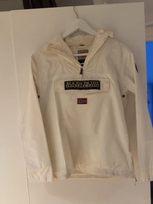 Napapijri - Snygg vit vindjacka från Napapijri med halv dragkedja, stor ficka framtill och norsk flagga som detalj. Jackan har huva och elastiska muddar vid ärmslut och nederkant. Perfekt för blåsiga dagar och streetwear-stil. Storlek S 