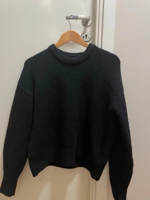 Svart stickad tröja med rund hals - En enkel och stilren svart stickad tröja med rund halsringning och långa ärmar. Tröjan har en något boxig passform och ribbstickade muddar vid ärmslut och nederkant. Perfekt att matcha med jeans eller kjol för en avslappnad look.