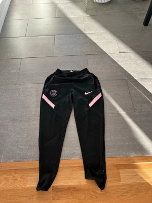 Svarta PSG träningsbyxor Nike - Säljer ett par svarta träningsbyxor från Nike med PSG-logga på ena benet och rosa detaljer. Byxorna har smal passform, resår i midjan och är tillverkade i ett lätt och stretchigt material som passar perfekt till träning. Storlek S men passar M 