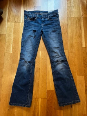Blå bootcut jeans med slitningar - Snygga blå bootcut jeans från Diesel med slitningar och hål på benen för en cool, avslappnad look. Klassisk femficksmodell och låg midja. Perfekta för dig som gillar en trendig och lite edgy stil. Dock har de ett hål i rumpan.