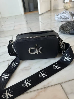 Svart axelväska från Calvin Klein - Snygg svart axelväska från Calvin Klein med silverfärgad CK-logga framtill. Väskan har två dragkedjefack och en bred axelrem med Calvin Klein-tryck. Tillverkad i slitstarkt material med en praktisk ficka på baksidan. Perfekt för att bära det viktigaste.