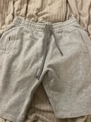 Grå mjukisshorts från H&M Divided - Gråa mjukisshorts från H&M Divided med elastisk midja och snörning. Shortsen har två sidofickor och är tillverkade i mjukt bomullsmaterial, perfekta för en avslappnad stil. Klassisk och enkel design som funkar till det mesta.